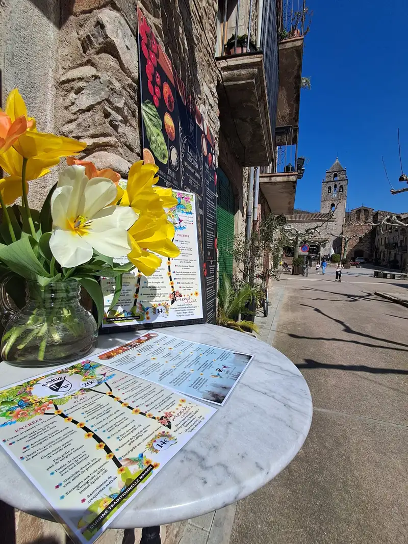 Mesa redonda de terraza con las cartas del Restaurant Samuga y jarrón de flores, frente a la plaza medieval de Sant Llorenç de la Muga en Girona