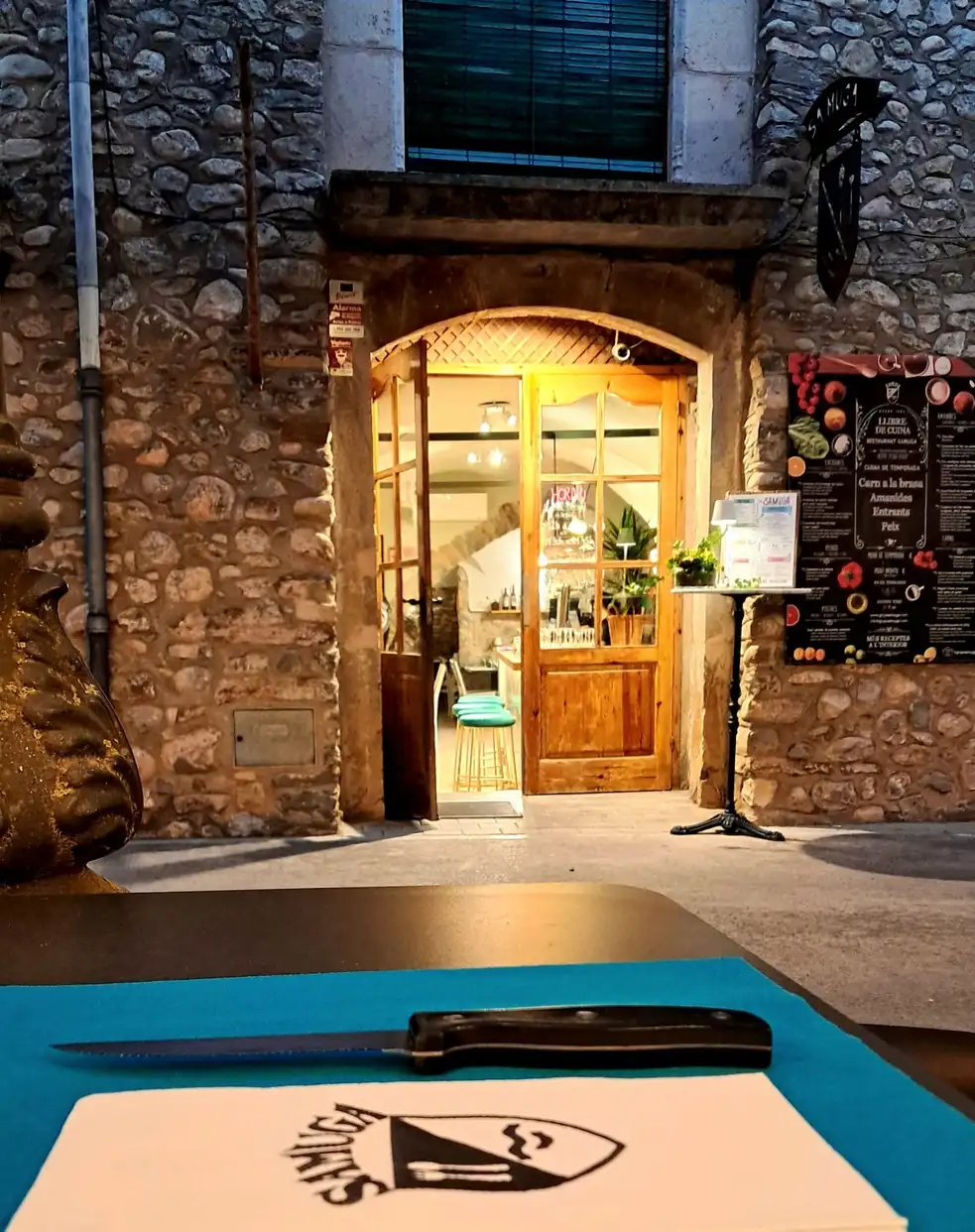 Entrada iluminada de Restaurant Samuga en la plaza medieval de Sant Llorenç de la Muga, Girona, vista desde mesa exterior con carta y cuchillo