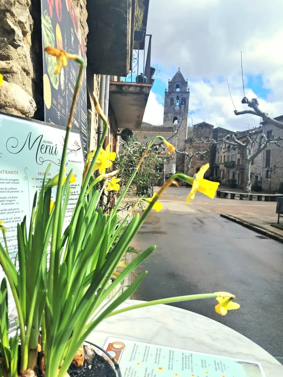 Terraza del Restaurant Samuga en Sant Llorenç de la Muga con cartas de menú, flores amarillas y vista al campanario del pueblo
