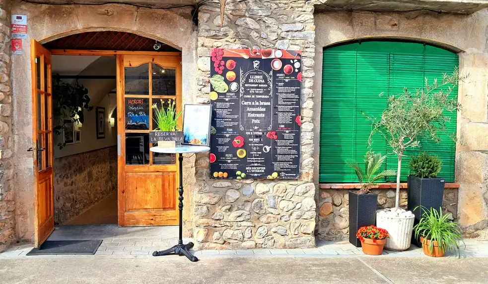 Fachada de piedra del Restaurant Samuga en Sant Llorenç de la Muga, Girona, con puerta de madera abierta y carta exterior en la pared