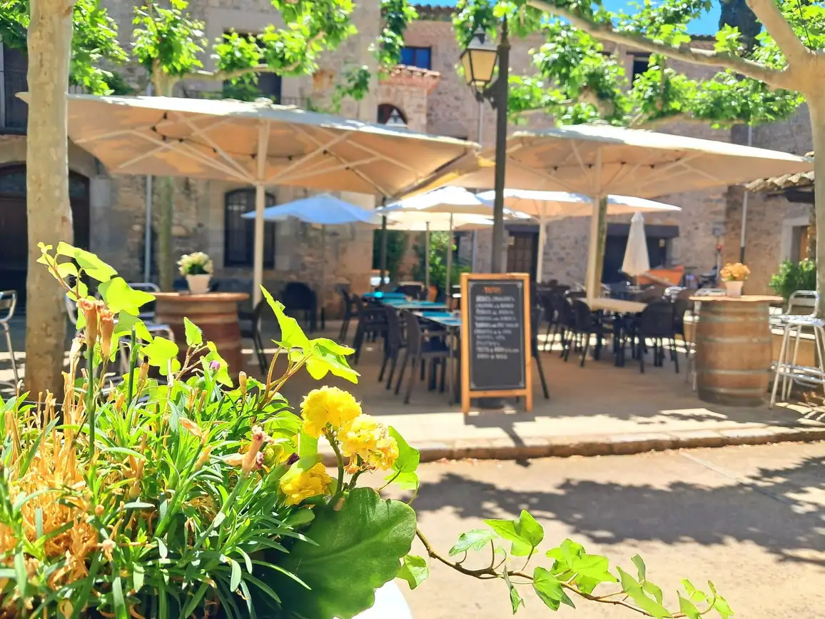 Terraza del Restaurant Samuga en la plaza medieval arbolada de Sant Llorenç de la Muga, Girona, con mesas al aire libre y pizarra de menú del día